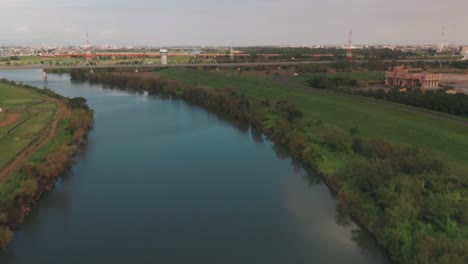 Drohne-Fliegt-über-Das-Ruhige-Wasser-Des-Arakawa-flusses-Mit-Einer-Eisenbahnbrücke-In-Der-Ferne