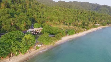 Tropical-Island-drone-backwards-dolly-shot-of-beach-resort-with-lush-green-rain-forest-and-tropical-palm-trees-with-white-sand-beach-and-rocky-coastline