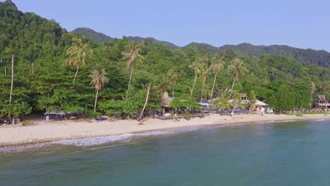 Tropical-Island-drone-backwards-dolly-shot-of-a-beach-resort-with-lush-green-rain-forest-and-tropical-palm-trees-with-white-sand-beach-and-rocky-coastline