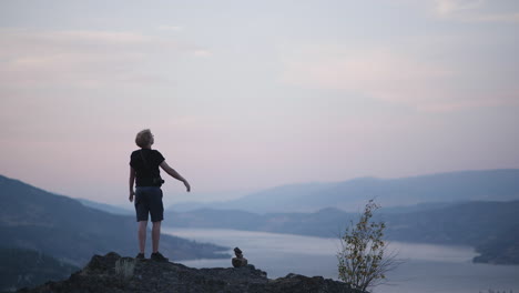 Hiker-stands-with-arms-raised-on-mountaintop-with-beautiful-sunset-view