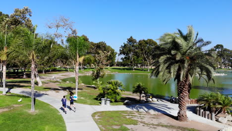 Fly-in-dolly-behind-igh-school-cross-country-runners-at-lake-at-Mason-Regional-Park-in-Irvine,-CA-shot-by-aerial-4k-drone