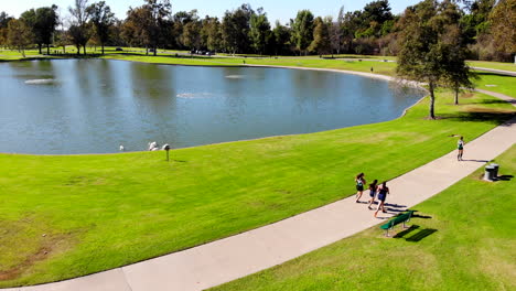 Girls'-high-school-cross-country-runners-pass-by-lake-at-Mason-Regional-Park-in-Irvine,-CA-shot-be-aerial-4k-drone
