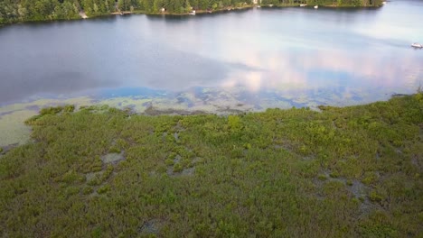 4k-aerial-clip-flying-from-land-over-a-canal-and-wetlands-to-the-lake-with-blue-water-and-green-trees-in-sunny-summer-conditions,-in-mid-michigan-on-Cranberry-Lake,-near-Harrison-MI