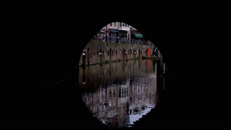 View-from-a-boat-peacefully-cruising-down-a-canal-in-Utrecht,-Netherlands:-moving-through-a-dark-tunnel-under-a-bridge,-seeing-the-cityscape-reflecting-in-the-water-and-panning-up-to-the-skyline