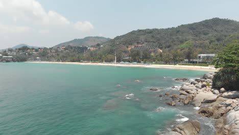 Rocky-shoreline-Cliff-splitting-Had-Karon-Noi-Beach-from-Karon-Beach-in-Phuket,-Thailand---Aerial-Low-angle-Tracking-shot-Reveal