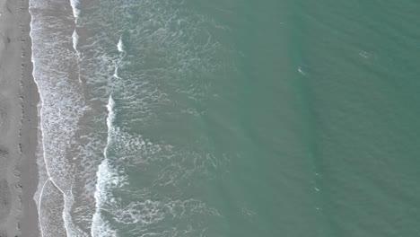 Eine-Luftaufnahme-Aus-Der-Vogelperspektive-Von-Meereswellen,-Die-Sanft-Am-Sandstrand-Aufsetzen-Und-Schaumstreifen-Verursachen