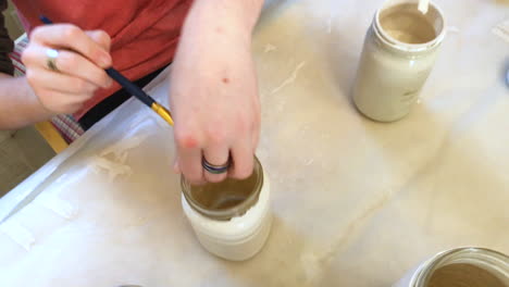 Teenage-boy-sitting-on-table-painting-mason-jars-with-white-paint,-top-view-and-camera-going-down-closer-to-action