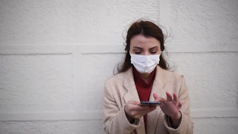 Slow-Motion:-Beautiful-girl-wearing-protective-medical-mask-and-fashionable-clothes-speaks-with-a-smart-phone-in-front-of-wall