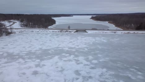 4k-Aerial-video-of-a-small-town-rural-frozen-lake-in-Michigan-in-the-winter-time,-small-bridge,-trees,-on-a-cold-overcast-day