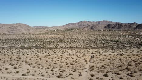 Cinematic-aerial-shot-of-desert-with-low-vegetation,-mountains-in-the-background-and-blue-sky