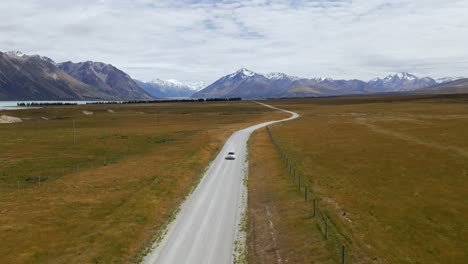 Aerial---car-driving-on-picturesque-dirt-road-with-snow-covered-mountains-in-background