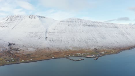 Flying-towards-Small-harbor-Town-at-sea-in-Westfjords,-Iceland-with-Boat-Dock---magnificent-White-Snow-Covered-Mountain-Ocean-Cliffs,-Drone-Aerial-over-arctic-waters
