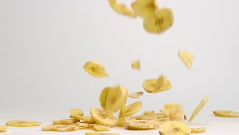 Crispy-yellow-freeze-dried-banana-chips-falling-down,-spinning-and-bouncing-into-a-pile-on-white-table-top-in-slow-motion