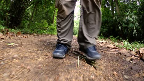 A-man-walking-in-the-jungle,-camera-focused-on-his-legs,-showing-him-stepping-forward-on-the-dirt-road-with-his-shoes-framed