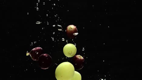 Red-and-green-grapes-dropped-into-water-with-floating-air-bubbles,-black-background