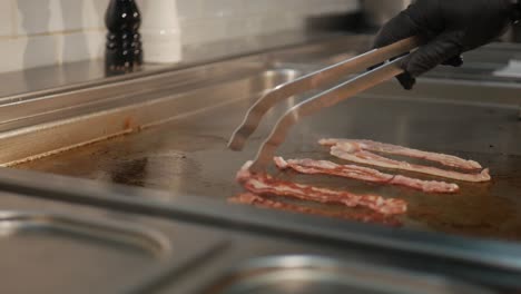 Chef-flip-over-tasty-bacon-strips-on-hot-stove-in-restaurant-kitchen,-close-up-shot
