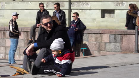 Father-and-Son-Relaxes-on-a-Stone-Pavement-with-Group-of-Friends-Resting-in-the-Background-on-a-Vilnius-Catheadral-Square