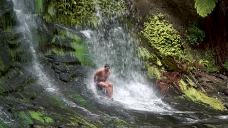 SLOWMO---Young-man-sitting-under-Mokoroa-Falls,-Auckland,-New-Zealand