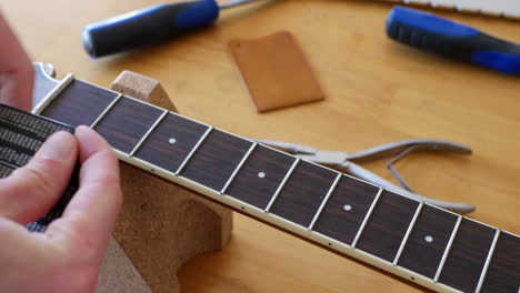Close-up-hands-of-a-luthier-craftsman-measuring-and-leveling-an-acoustic-guitar-neck-fretboard-on-a-wood-workshop-bench-with-lutherie-tools-SLIDE