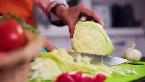 A-slow-mo-side-angle-close-up-view-of-a-lady-chef-cutting-cabbage-with-knife-in-the-kitchen,-A-basket-of-veggies-with-tomatoes-and-garlic-on-the-table,-The-chef-wore-and-orange-colored-apron