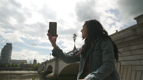 Hispanic-Latina-tourist-using-Skype-and-showing-her-friend-the-river-Thames-and-Putney-Bridge-in-London,-smiling-and-having-a-great-time