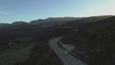 Following-white-car-driving-curve-on-mountain-road-with-beautiful-wide-far-cinematic-landscape-scenerie-and-forrest---Aerial-helicopter-drone-shot