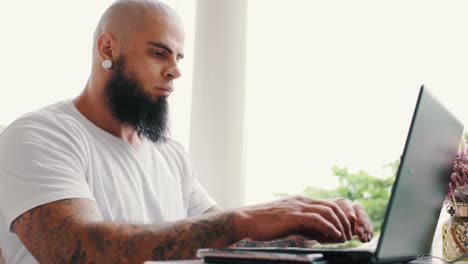Handsome-brutal-young-man-with-beard-and-tattoos-is-working-on-laptop-near-window-in-bright-space