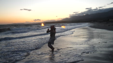 A-man-fire-dancer-and-performer-in-silhouette-spinning-his-flaming-staff-in-the-ocean-water-on-the-beach-at-sunset-in-slow-motion