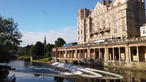 Static-Shot-of-Pulteney-Weir-and-the-Empire-Hotel-in-Bath,-Somerset-on-Beautiful-Summer’s-Morning-with-Blue-Sky-with-Birds-Flying-Into-Frame