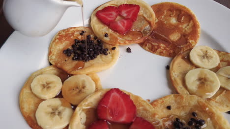 Pouring-Maple-Syrup-in-a-circular-motion-over-an-elegant-arrangement-of-pancakes-with-sliced-strawberries,-banana,-and-chocolate
