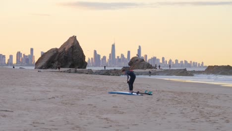 Surfer-In-Wetsuit-Changing-Surfboard-In-Currumbin-Alley-During-Sunset---Surfers-Paradise-Skyscrapers-On-The-Background---Gold-Coast,-Queensland,-Australia