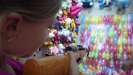 Junges-Kaukasisches-Mädchen,-Das-Gummigewehr-Auf-Reihen-Bunter-Luftballons-Schießt,-Um-Preise-Am-Schießstand-Zu-Gewinnen