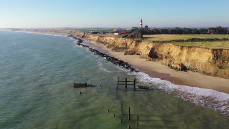 Moving-footage-towards-the-lighthouse-at-happisburgh,-Norfolk,-UK-by-drone-from-the-sea-defences-with-the-tide-coming-in-in-the-summer