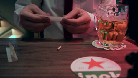 Man-Rolling-Cannabis-Joint-On-A-Bar-Table-With-A-Glass-Of-Beer-In-Amsterdam,-Netherlands---close-up