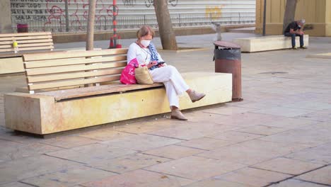 Adult-Woman-Wearing-Face-Mask-Sitting-On-The-Wooden-Bench-And-Browsing-On-Her-Mobile-Phone-In-Plaza-de-la-Merced-During-The-Pandemic-Coronavirus-In-Malaga,-Spain