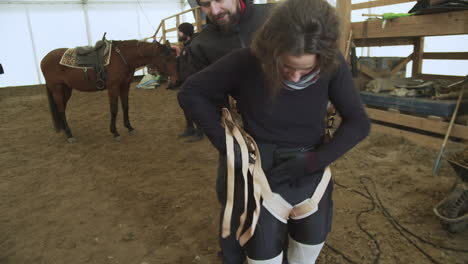 Stuntwoman-adjusts-harness-on-legs-with-horse-and-stunt-crew-in-background
