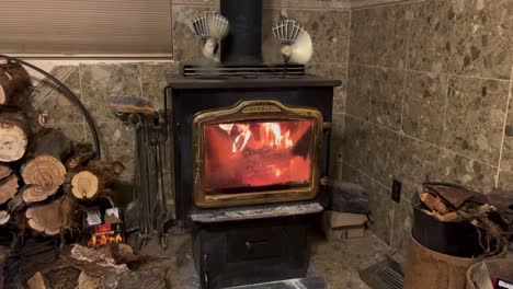 A-wood-burning-stove-with-firewood-stacked-next-to-it-burns-strong-with-two-kinetic-fans-sitting-on-top-of-the-stove