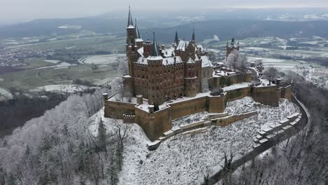 Aerial-flying-towards-and-panning-down-medieval-Castle-Hohenzollern-on-a-mountain-on-a-snowy-day-during-winter-in-Swabia,-Germany