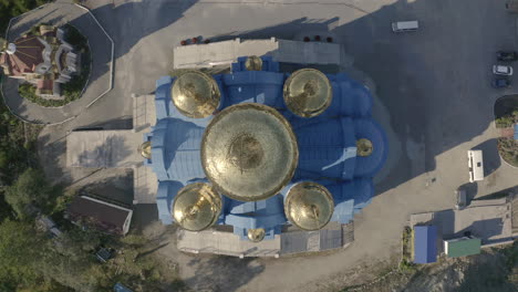 Slow-descent-90-degree-aerial-shot-of-an-orthodox-church-with-blue-roof-and-golden-domes,-located-on-top-of-the-hill-on-a-bright,-clear,-sunny-day