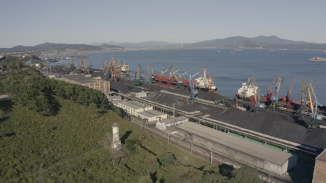 Aerial-shot-of-a-coal-port-with-moored-general-cargo-bulk-carrier-ships-and-a-bay-with-green-hill-side,-on-a-bright-sunny-day