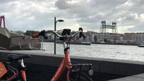 Orange-Donkey-Republic-Bicycle-parked-in-front-of-Rotterdam-port-with-cargo-vessel-sailing-through-waterway