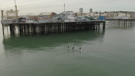 Drei-Leute-Auf-Paddelbrettern,-Die-Am-Brighton-Pier-Vorbei-Zum-Strand-Auf-Grünem-Wasser-Gehen