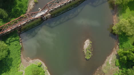 Aerial-drone-flying-above-reflective-blue-river-and-old-rusty-truss-bridge-alongside-a-bright-green-forest-in-central-Pennsylvania-during-the-summer