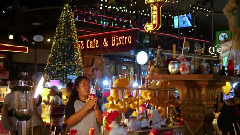 Mujer-Rezando-En-El-Altar-Budista-En-El-Mercado-Nocturno,-Turistas-Que-Pasan-Por-Bangkok