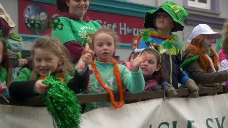 Group-of-children-stand-in-truck-wagon-dressed-in-green,-wearing-Irish-symbols,-clovers-and-green-hats,-waving-to-camera-and-smiling