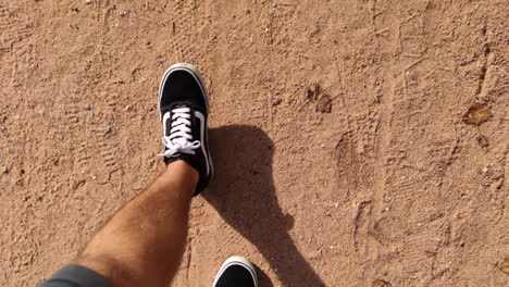Slow-Motion-Of-A-Man's-Feet-In-Black-Vans-Sneakers-Walking-On-Sandy-Ground-On-A-Sunny-Day---overhead-shot,-POV