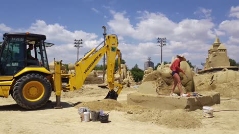 Excavator-At-Work-At-Sand-Festival-In-Burgas,-Bulgaria-With-Man-Watering-Artistic-Sculpture-Made-Of-Sand---wide-shot