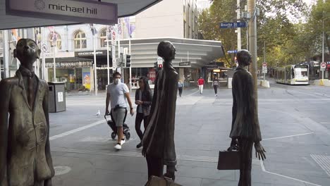 People-wearing-masks-walk-through-Melbourne's-quiet-streets,-passing-popular-statues-during-the-coronavirus-COVID-19-lockdown---Melbourne,-Australia
