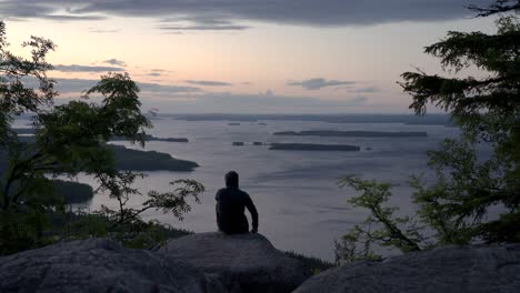Hooded-male-hiker-sitting-on-a-nordic-mountain-cliff-looking-towards-horizon-and-enjoying-a-summer-sunset-or-a-sunrise-over-a-big-lake-view