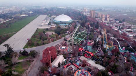 Aerial-orbit-of-amusement-park-extreme-rides-and-ellipse-music-dome-venue-in-O’Higgins-Park,-Santiago-city-in-background,-Chile